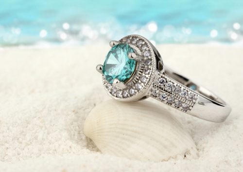 An aquamarine ring leaning against a seashell on a beach