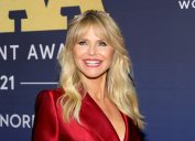 Christie Brinkley in front of a navy blue background smiling and wearing a red satin blazer