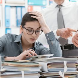 Demanding boss pointing to his watch and asking his employee to hurry up while she sits behind a stack of folders and papers