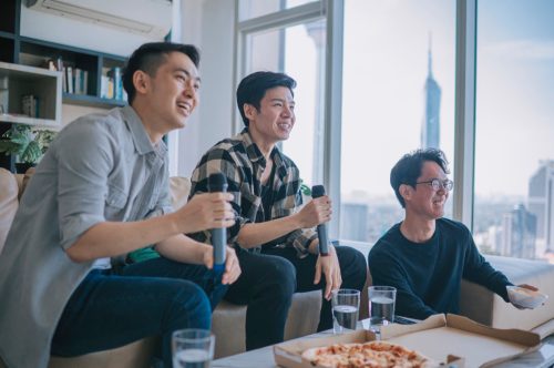 male friends enjoying karaoke session at home during weekend eating pizza