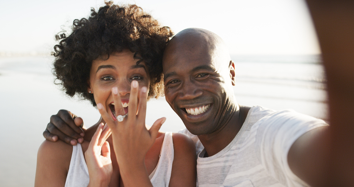 Cropped photo of a happy young couple taking a selfie on the beach, showing off their engagement ring.