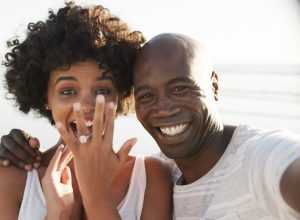 Cropped photo of a happy young couple taking a selfie on the beach, showing off their engagement ring.
