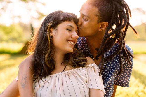 Beautiful young interracial couple being romantic while sitting in a park. Affectionate young man kissing his girlfriend on her forehead while embracing her during the day.