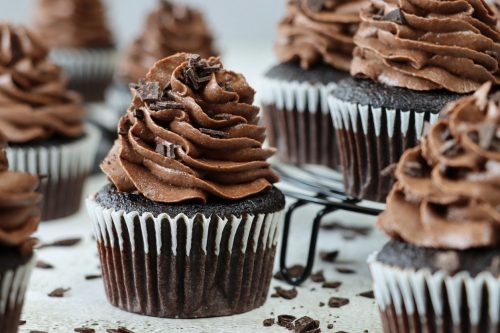 freshly baked, homemade, chocolate cupcakes in paper cake cases on a circular, black, metal wire cooling rack. The cup cakes have been decorated with swirls of chocolate piped icing and chocolate pieces.