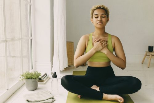 Young African American lady practicing yoga at home in black leggings sitting on floor in lotus posture hands crossed on chest slight delight smile on face, feeling her body. Emotional health