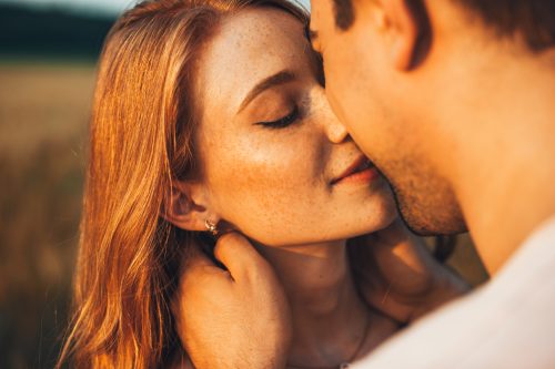 Close-up portrait of a freckled girl kissing her boyfriend while they are on an outdoor date. Wheat field.