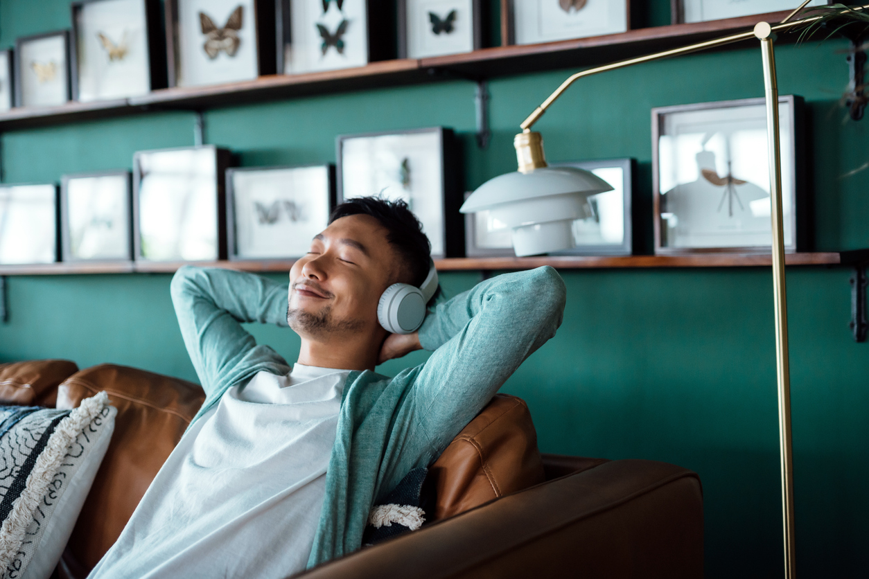 Young Asian man with hands behind head, relaxing on sofa and listening to music with headphones at home. Relaxed young man lying on sofa with music.