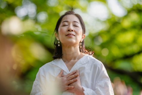 A woman is closing her eyes, doing breathing exercise and meditating in nature.
