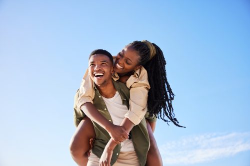Laughing young African man carrying his smiling girlfriend on his back while enjoying a day at the beach together on a sunny day in summer