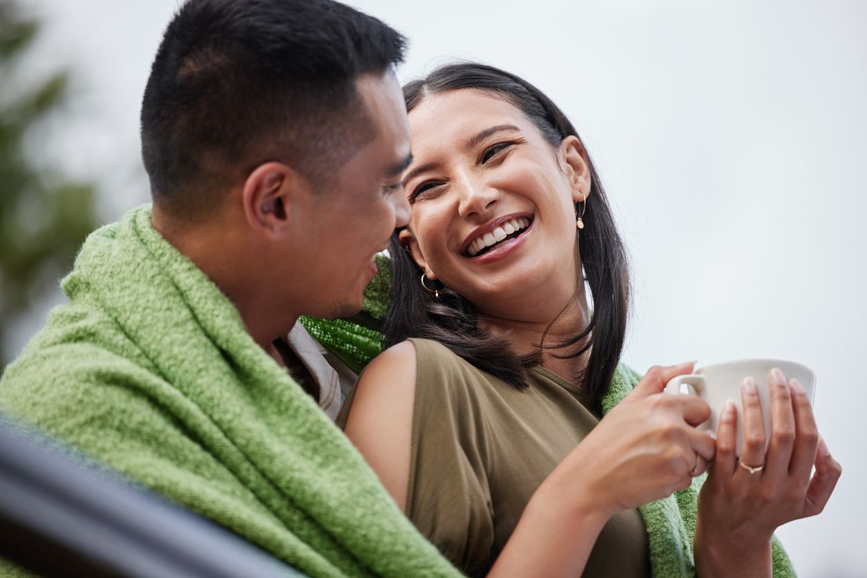 Cozy, smiling and romantic young couple cuddling and relaxing with blanket in cold winter. Beautiful, loving husband and wife laughing and enjoying dating relationship bonding time in outdoors nature