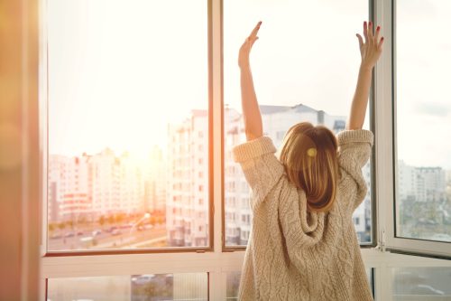 Woman near window raising hands feeling refreshed after receiving a good morning text message