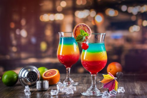 Colorful layered tropical cocktails on a bar surrounded by fruit