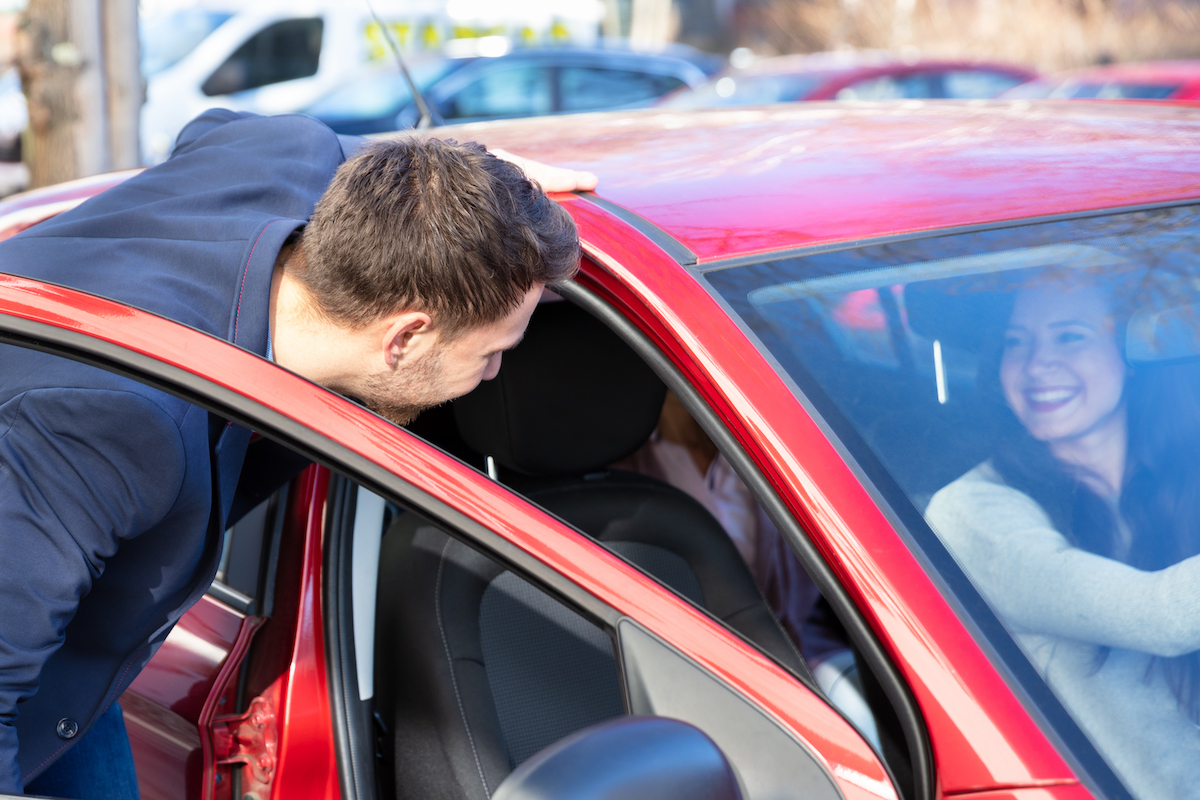 A smiling man getting into a red car with a female in the passenger seat.