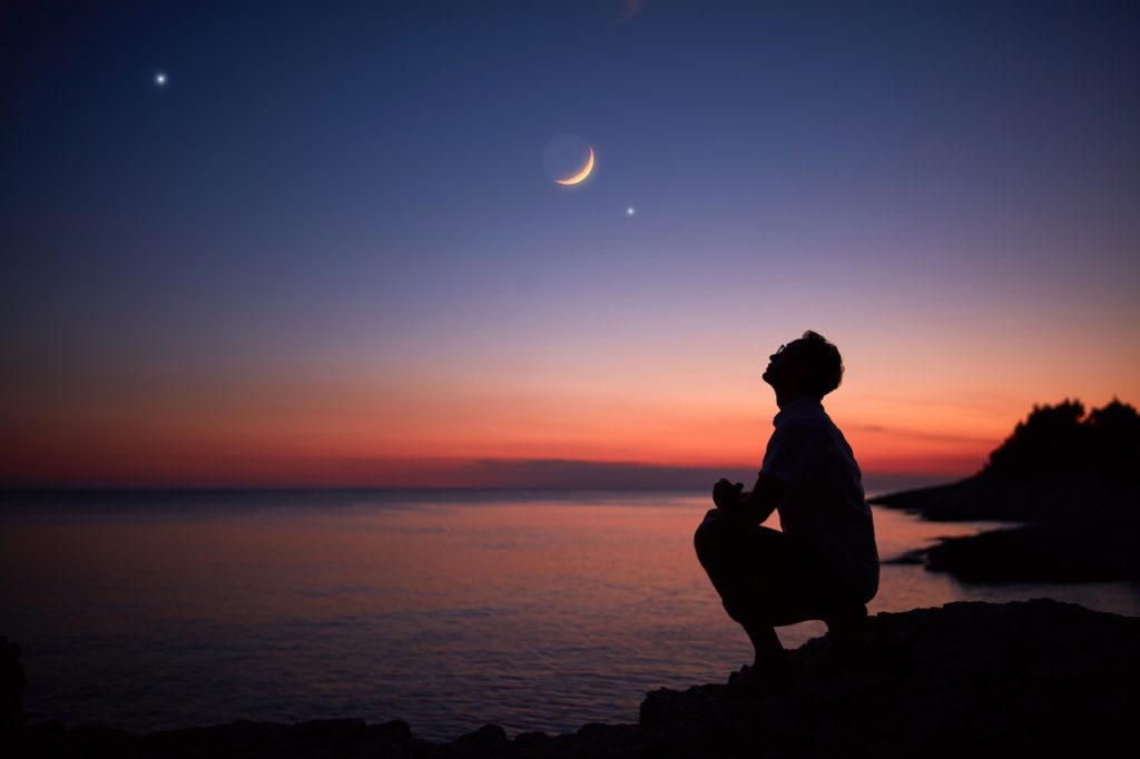 Hình bóng của một người đứng bên đại dương và nhìn mặt trăng và các hành tinh trên bầu trời đêm lúc chạng vạng