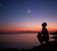 The silhouette of someone standing by the ocean and looking up at the moon and planets in the night sky at dusk