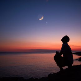 The silhouette of someone standing by the ocean and looking up at the moon and planets in the night sky at dusk