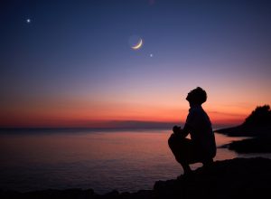 The silhouette of someone standing by the ocean and looking up at the moon and planets in the night sky at dusk