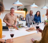 Group Of Mature Friends Meeting At Home Preparing Meal And Drinking Wine Together
