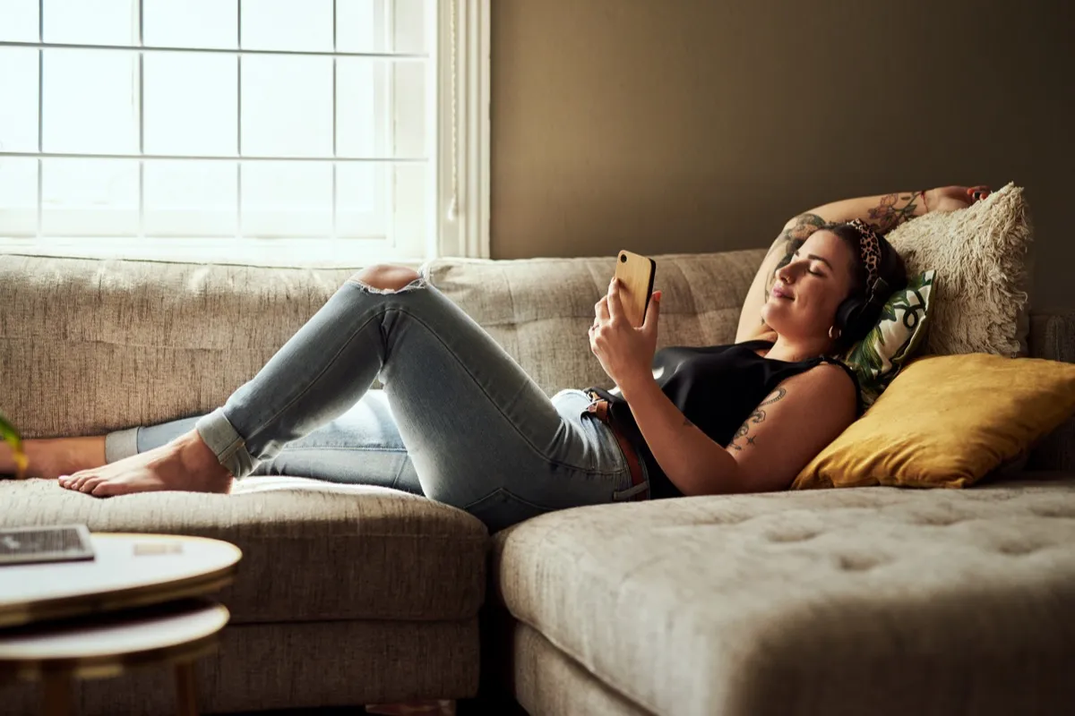 Shot of a young woman using a smartphone and headphones on the sofa at home