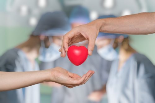 hand passing a red heart while doctors perform organ transplant in background