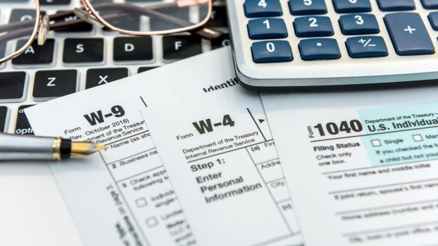 A stack of tax forms on top of a keyboard next to a pair of glasses and a calculator