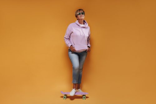 Full length photo of cool woman with short hair in sunglasses, wide hoodie and skinny jeans smiling and posing with pink skateboard..