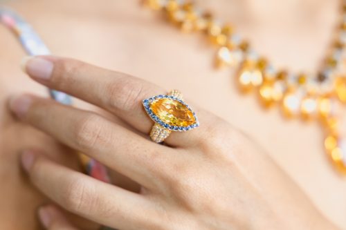 Close up of a big topaz and diamond ring on a woman's hand, with a matching necklace visible in the background