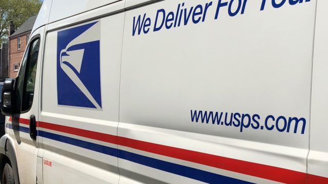 Angled closeup of the side of a USPS van (with logo, tagline, and website address) parked in Queens, NY