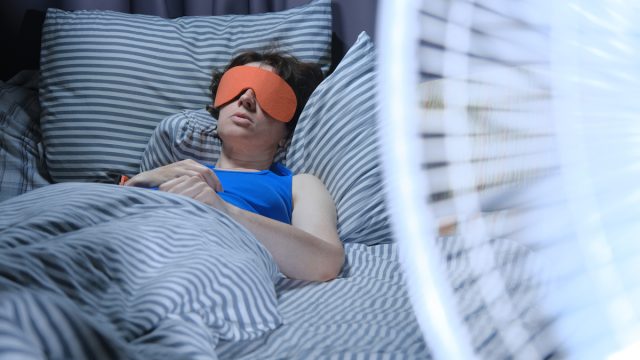Woman is using eye mask in bed while electric fan is blowing on the foreground