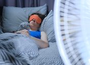 Một người phụ nữ sử dụng mặt nạ mắt trên giường trong khi một chiếc quạt điện thổi ở phía trước