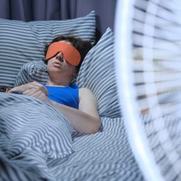 Woman is using eye mask in bed while electric fan is blowing on the foreground
