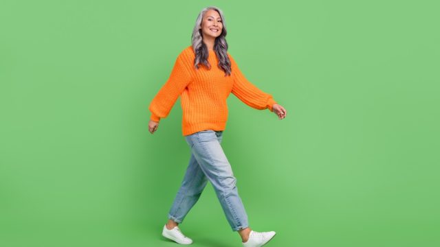 Full body photo of woman with gray hair wearing slouchy boyfriend jeans and sneakers isolated on green color background