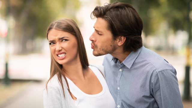 A woman with a disgusted face pulling away from a man trying to kiss her while outside in a park