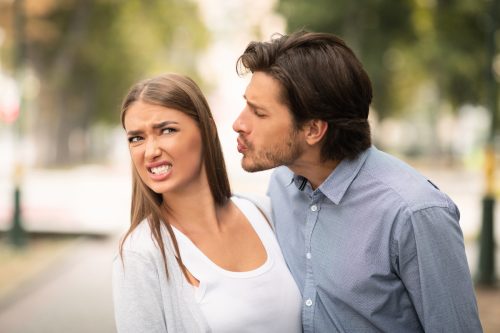A woman with a disgusted face pulling away from a man trying to kiss her while outside in a park