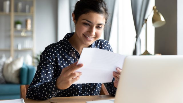 A young woman smiling at a letter she received while sitting in front of her laptop