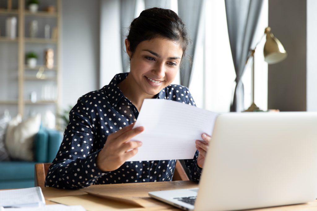 A young woman smiling at a letter she received while sitting in front of her laptop