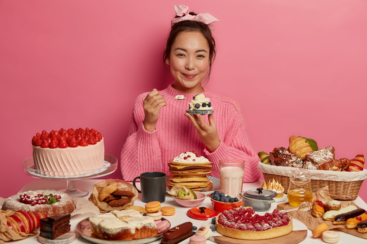 A smiling young woman wearing a pink sweater, sitting behind a table of desserts, with a pink background.