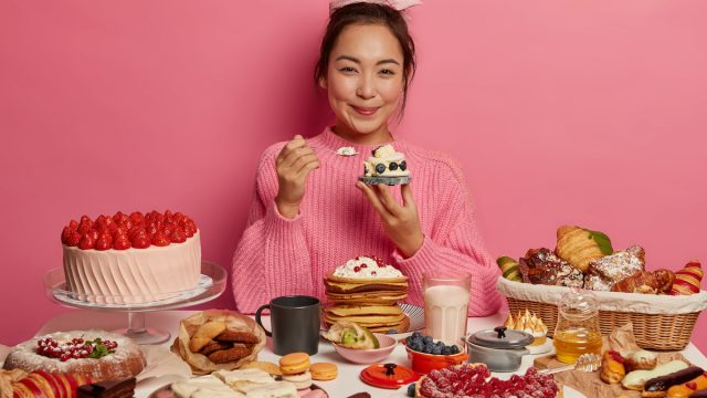 A smiling young woman wearing a pink sweater, sitting behind a table of desserts, with a pink background.