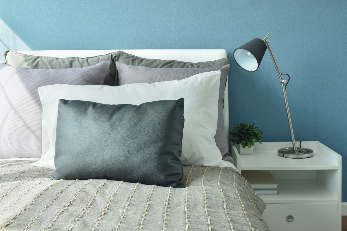 Gối màu xám, xám đậm và trắng trên giường với đèn bàn kiểu hiện đại và tường màu xanh