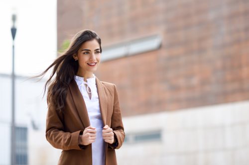 A smiling woman with long brown hair wearing a white shirt and brown blazer outside.