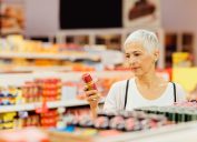 Người phụ nữ trưởng thành mỉm cười mua sắm tại siêu thị địa phương.  Đứng cạnh quầy sản phẩm và chọn gia vị.  Đọc nhãn dinh dưỡng.