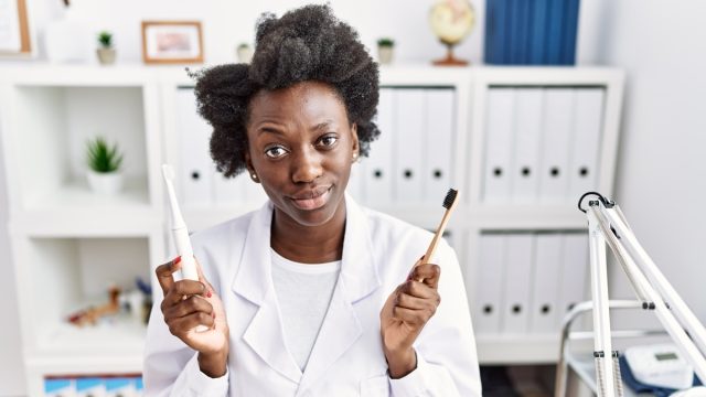 African dentist woman holding electric toothbrush and normal toothbrush skeptic and nervous, frowning upset because of problem.