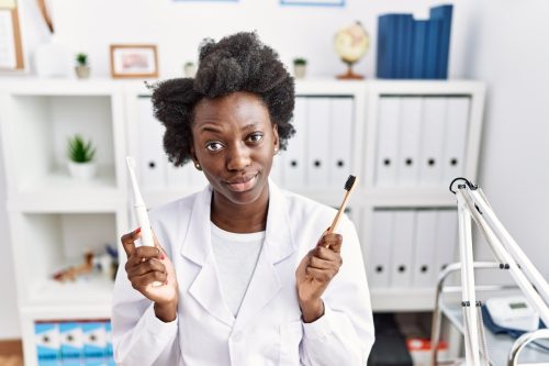 African dentist woman holding electric toothbrush and normal toothbrush skeptic and nervous, frowning upset because of problem.