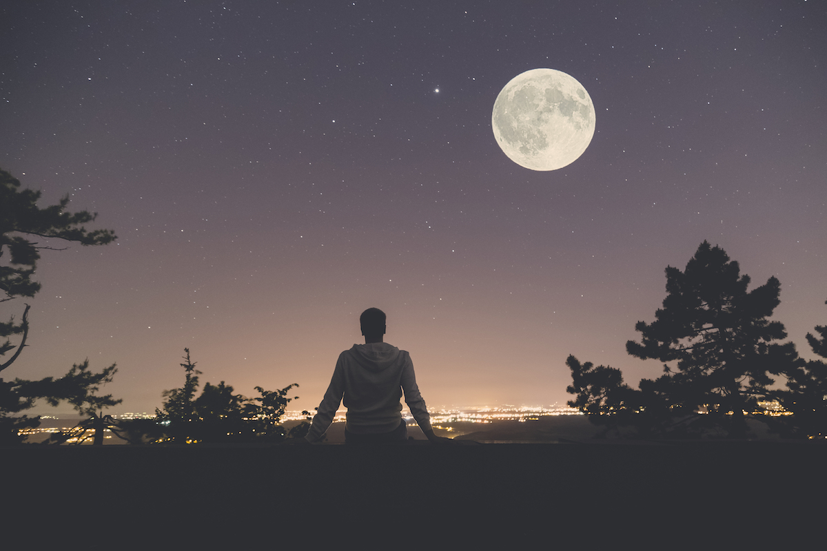 Young man sitting on the wall at night. City lights, moon and stars in the background.