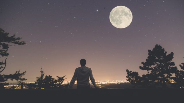 Young man sitting on the wall at night. City lights, moon and stars in the background.