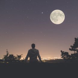 Young man sitting on the wall at night. City lights, moon and stars in the background.