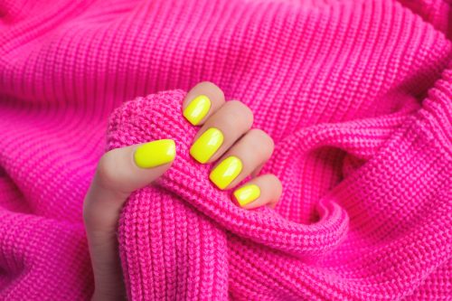 Một bàn tay có móng tay màu vàng neon cầm chiếc áo len màu hồng tươi