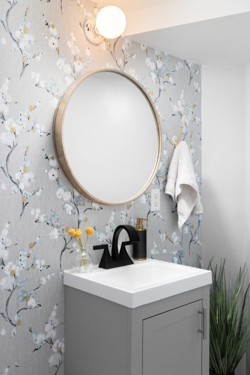 Detail of a bathroom with a grey cabinet, wooden circular mirror, and patterned wallpaper.