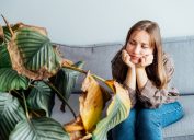 A young woman sitting on her couch looking sad and staring at a big, dead houseplant.