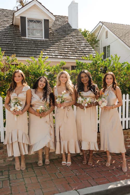Models standing in front of a house with a white picket fence wearing champagne-colored satin bridesmaid dresses from Show Me Your Mumu